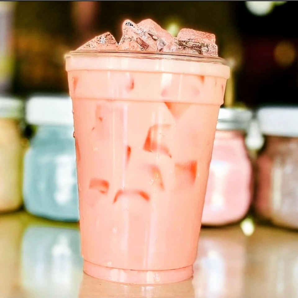 Pink iced drink in a clear plastic cup, with colorful jars blurred in the background.