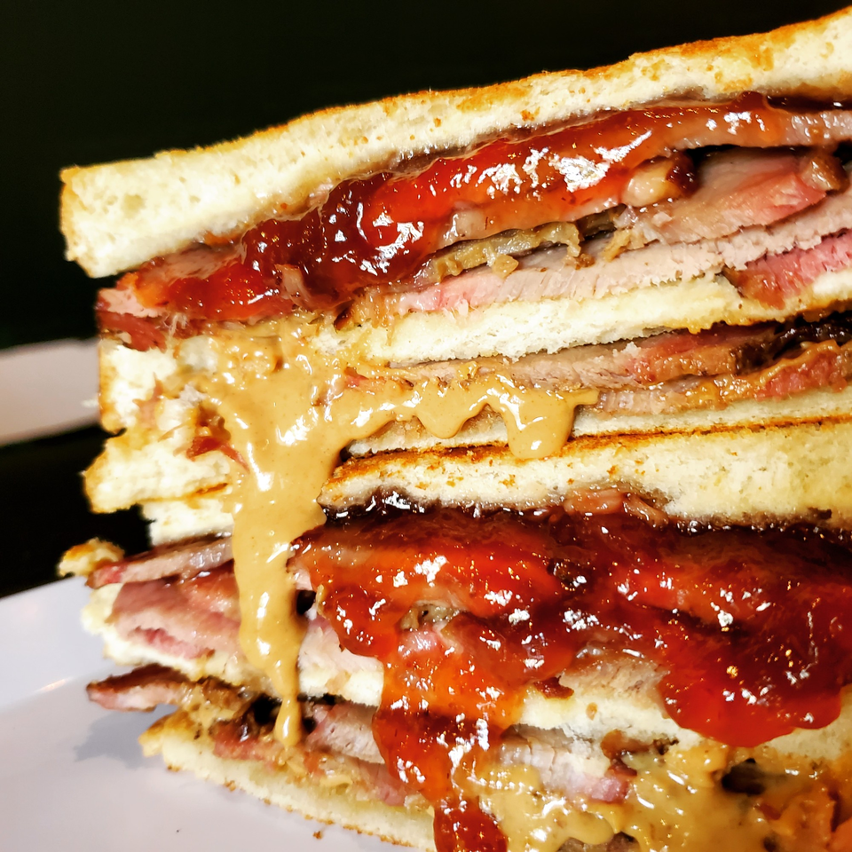 A close-up of a multi-layered sandwich with meat, peanut butter, and jelly oozing out.