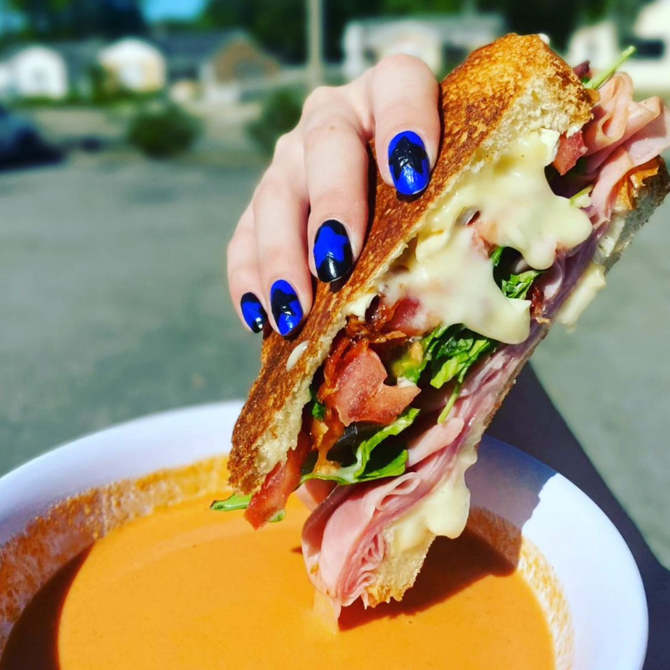 A hand with blue and black nails holds a grilled sandwich over a bowl of tomato soup.