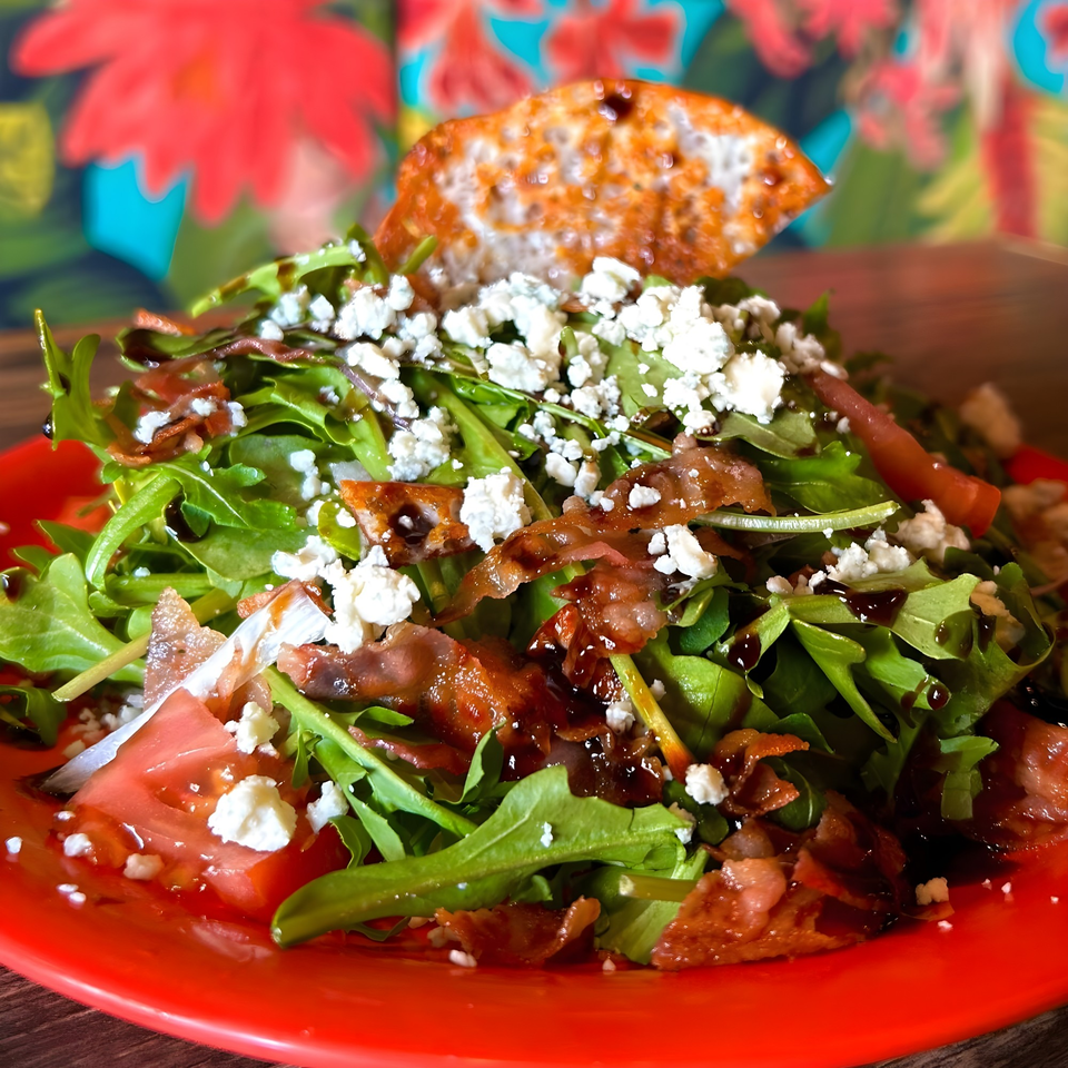 Arugula salad with crumbled feta, bacon, tomatoes, and balsamic glaze on a red plate.