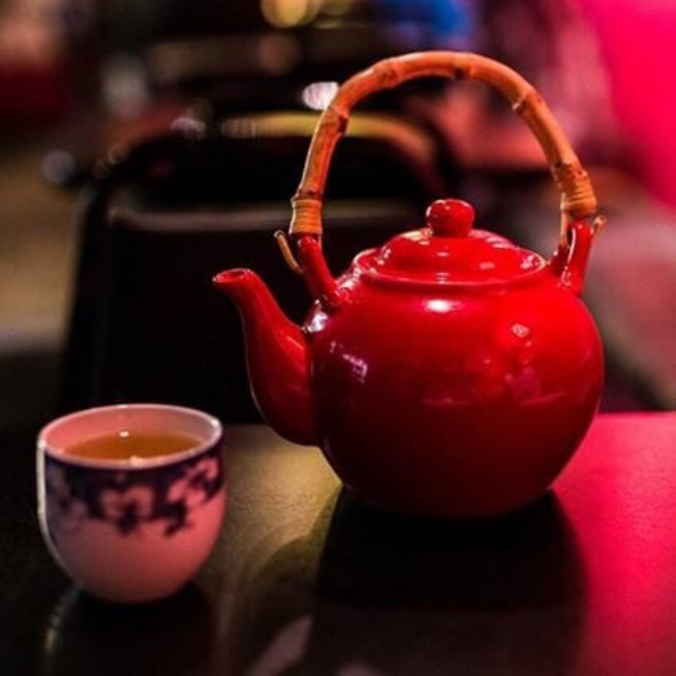 A red teapot with a bamboo handle sits beside a small cup of tea on a dark surface.